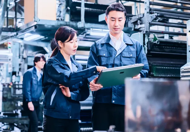 機械が並ぶ職場で、二人で協力して仕事を進める様子。